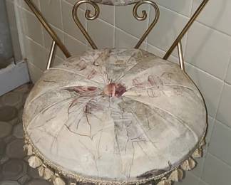 A vintage round-back accent chair with a light floral upholstered seat and backrest, trimmed with a full ring of tassels around the cushion. The metal frame has a gold/brass finish with decorative scrollwork on the back supports and curved legs connected by ornate metalwork at the base. The upholstery shows visible age and wear, giving it a shabby‑chic, romantic, or Victorian‑inspired look. It’s the kind of piece people use as a vanity chair, boudoir accent, or decorative seating.  Priced at $100