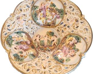 A Capodimonte decorative ornate porcelain display plate featuring raised classical‑style scenes in high relief. The center shows three standing figures in a mythological or allegorical setting, while the two side panels depict reclining figures with animals and lush foliage, all sculpted with fine detail. The rim is pierced with swirling openwork and finished with gold accents, giving the plate an elegant European look—similar to Rococo or Neoclassical revival pieces. The craftsmanship and gilding make it a striking wall or cabinet display piece.  Priced at $350