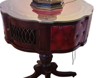 A round vintage side table with a scalloped glass‑top surface and an ornate gold inlay border. The body combines dark wood with red tufted leather‑style panels and a section of decorative metal latticework, giving it the look of a mid‑century or early‑20th‑century accent piece—possibly from a line of furniture that incorporated speaker grilles or decorative vents into the design. It stands on a central pedestal base with four curved legs ending in brass claw‑style feet, adding to its antique character. The lamp on top complements the table’s ornate style, but the pricing below reflects the table only.   Priced at $200