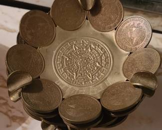 A small decorative Mexican coin ashtrays crafted from authentic old pesos, arranged in a circular pattern to form the rim. Each coin shows the national emblem of Mexico—an eagle perched on a cactus with a snake in its beak—giving the piece strong cultural character. The center features a raised Aztec Sun Stone (Calendar) design, adding depth and historical symbolism. The dish has a handcrafted folk‑art feel and works well as a display piece, catch‑all, or collectible item.  Priced at $10 ea