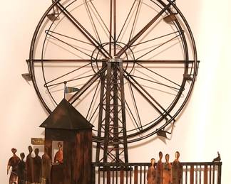 A metal wall sculpture depicting a Ferris wheel and carnival scene, crafted from thin metal rods and flat cut‑metal figures. The Ferris wheel is the centerpiece, with a circular frame, radiating spokes, and small gondolas attached around the rim. In front of it sits a ticket booth topped with a tiny flag and a “Tickets” sign, adding charm and context. Several stylized human figures stand near the booth and along a fence-like rail, giving the artwork a sense of movement and storytelling. The mixed metallic tones and handcrafted construction give it a rustic, whimsical, and nostalgic feel—perfect for eclectic, industrial, or vintage-inspired décor.  Price: $50