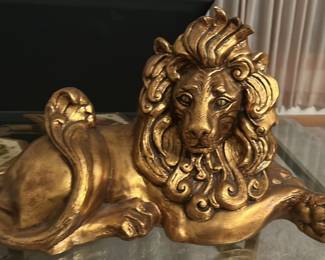 The lion is a large, decorative gold sculpture designed in a dramatic, regal style. It’s sculpted in a reclining pose, with one front paw extended forward and the head lifted proudly, giving it a commanding presence. The mane is the standout feature—full, flowing, and highly stylized, with deep curls and ridges that create a sense of movement and luxury.  The finish is a bright metallic gold, smooth and reflective, meant to catch light and draw attention. The body is elongated and elegant rather than anatomically realistic, which places it firmly in the world of Hollywood Regency or mid‑century decorative art—pieces made to be glamorous statement décor.  Priced at $250