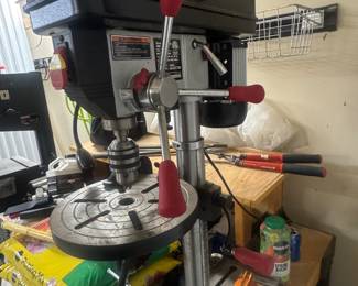 Bench-top drill press with red handles on a workbench, surrounded by tools and supplies.