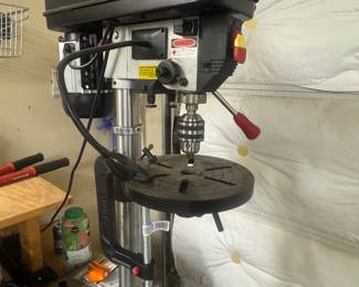 Standing drill press with adjustable platform and red handle, positioned against a wall in the garage.