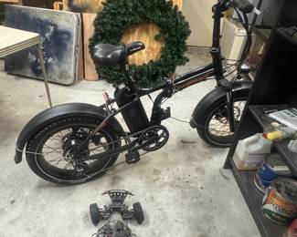 Black bicycle with a large green wreath hanging on the frame, plus a small toy car on the floor in a garage.