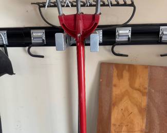 Red-handled yard tool (possibly a string trimmer) hanging on a wall rack along with wooden boards.
