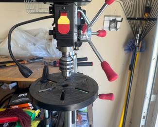 Craftsman bench-top drill press with red handles, surrounded by yard tools and equipment in the garage.