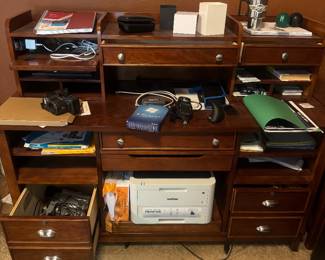 Wooden office desk with multiple drawers and compartments, cluttered with electronics, papers and desk supplies.