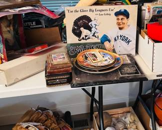 Vintage sports memorabilia table featuring baseball gloves, baseballs, bats, bobbleheads, trading cards, and baseball collectibles, including a Lou Gehrig advertising sign and mixed MLB decor. Great selection for sports collectors, baseball fans, and resellers.