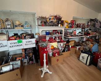 Wide room view featuring vinyl records, jukebox-style radios, gumball machines, Coca-Cola and beer advertising signs, sports memorabilia, framed collectibles, and mixed vintage display pieces. Great variety of music, advertising, pop culture, and nostalgia items throughout this packed picker-style sale.
