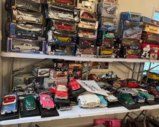 Large die-cast and model car collection featuring vintage cars, hot rods, motorcycles, classic trucks, and boxed collectible vehicles in multiple scales. Shelves and floor displays are packed with display-ready models, loose cars, and reseller-friendly automotive collectibles.