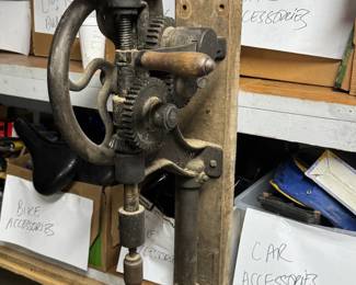 Antique wall-mounted hand-crank drill press with cast iron gears, mounted on original board, displayed in the garage/tool area near boxes of car and bike accessories. Great industrial piece for collectors of old tools, workshop decor, and vintage shop equipment.