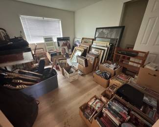 Overflow room with framed artwork, prints, mirrors, DVDs, and media boxes alongside guitars, instrument cases, and music-related pieces. Great mix of wall art, vintage media, and musician/reseller finds with plenty more to sort through.