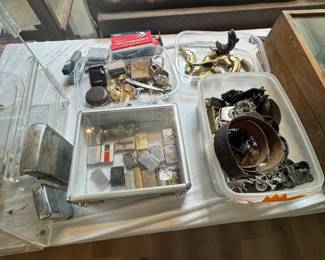 Table of vintage lighters, cigarette cases, jewelry, belt buckles, brass decor, and collectible men’s accessories, plus display cases filled with smalls. Great assortment of pocket-size vintage finds for collectors, resellers, and treasure hunters.