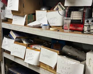 Organized garage shelf section with sorted boxes of heaters, lanterns, propane items, old lunch boxes, thermoses, bike helmets, car accessories, batteries, and battery chargers. Great picker-friendly setup with categorized garage lots, useful vintage storage finds, and resale inventory.