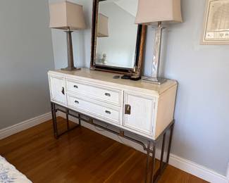 White chest on black stand with a black and gold mirror and a pair of silver lamps