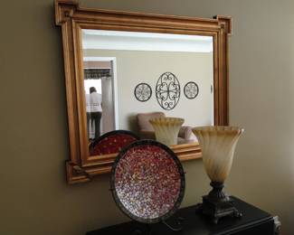 Gold framed mirror, with a decorative plate, and a nice lamp next to it.  Mirror mirror on the wall, can you see the metal artwork on the wall