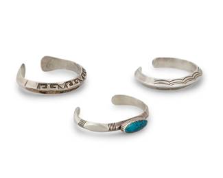 1384 A group of Southwest cast silver cuff bracelets Mid/late 20th century Three works:  Will Denetdale (b. 20th century, Diné) A Navajo sterling and turquoise cuff Stamped: W Denetdale / Navajo / Sterling [script, partial W] A cast silver cuff with narrow oval set turquoise cabochon and incised line accents 6.25" total inner C x 0.375" H, gap: 1.25"  Two Southwest cast silver cuff bracelets One stamped: RC / C [on flower within circle] Comprising a chunky cast silver ridged cuff with geometric and pictorial silver and 12K gold plate overlay, and a chunky silver ridged cuff with stamp work accents 2 pieces Each cuff: 6.125" total inner C x 0.375" H, gap: 0.75"  3 pieces total 126.4 grams gross Estimate: $600 - $900