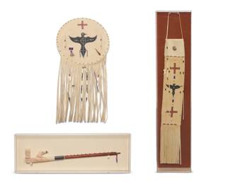 1319 John Redtail Freesoul (b. 20th century, Cheyenne-Arapaho) A group of Cheyenne-Arapaho custom-made painted hide items, 1987 & 1991 Three works:  A "Hawk Ceremonial" effigy pipe, 1987, with a carved alabaster bowl, and dark pipestone accent, on a wood stem with antique French brass tacks, and trade bead dangles, professionally mounted and framed under acrylic Signed to base of black stone 23.5" L x 3.5" H x 1.25" W approximately  A "Shadow of the Hawk" buckskin pipe bag, 1987, featuring glass trade bead beadwork, painted cross motifs, and a central bird in black, with long fringe and bone and bead dangles, professionally mounted and framed under acrylic 28" H 6.25" W; fringe: 13" approximately  A "Hawk Dream Shield", 1991, in bull hide featuring a central large painted black bird, and with feathers, and brass and trade bead accents, and bead and bone dangles within the long fringe Signed, titled, and dated verso 18.75" Dia. x 1" D, fringe: 18.75"  3 pieces Estimate: $800 - $1,200