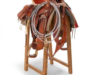 1081 A Juan Lara Western saddle with accessories Late 20th/21st century; Santa Barbara, CA Embossed to seat back and to front swell tab: Juan Lara / Santa Barbara / 2772 The smooth brown leather saddle with contrast stitching and braided leather lace accents, silver conchos with laces, skirt, fenders and multiple straps and rings, the stirrups and cantle trimmed with rawhide and the seat with sheepskin padding, seat size 16, together with a woven horsehair girth with decorative tassel, a pair of saddlebags, a wide martingale strap, a striped twill weave saddle blanket, a lariat, and two Faulks wooden duck calls, as well as a wooden saddle stand 8 pieces Seat: 16"; Swell width: 14"; Gullet width: 4"; Stand: 40.25" H x 16" - 21.25" W x 24" D Estimate: $500 - $700