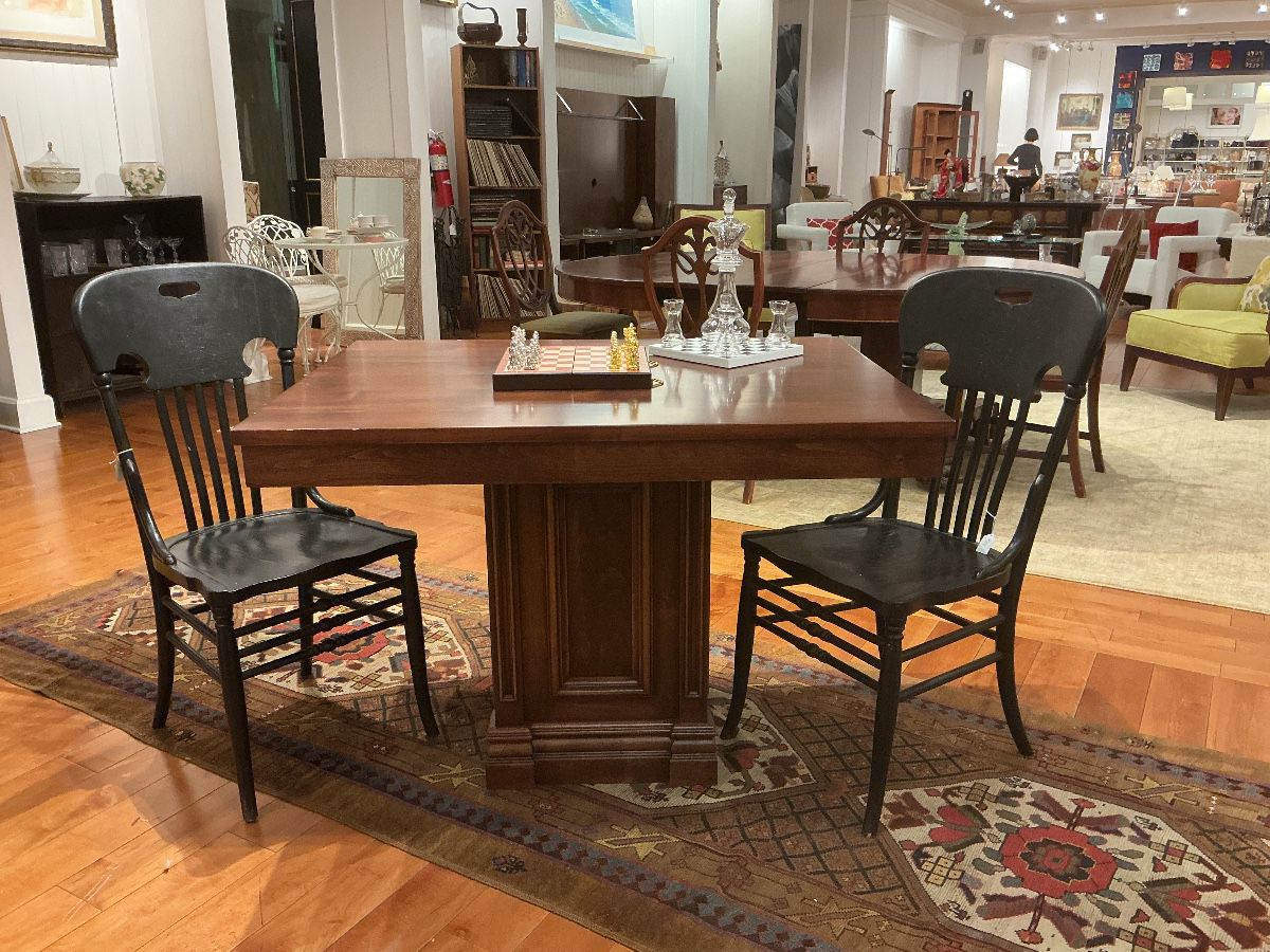 Heavy mahogany pedestal games table with a robust architectural base, paired with two vintage ebonized spindle-back chairs. The chairs feature "ox-yoke" top rails with integrated handles and a charming, factory-distressed finish. A perfect blend of traditional weight and rustic contrast.