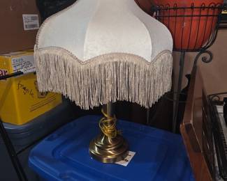 This item is a vintage-style table lamp featuring a cream-colored, scalloped lampshade with extensive fringe trim and a brass-finished base.