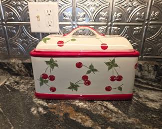 This item is a vintage Target Home red cherries ceramic bread box, originally retired around 2005. It is characterized by its white ceramic body decorated with red cherries and green leaves, finished with red trim and a matching lid with a handle.