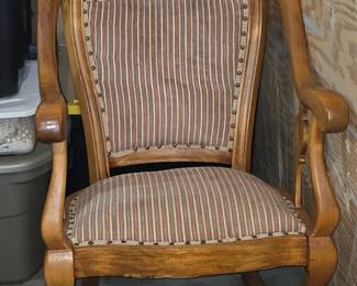 This item is a vintage upholstered rocking chair, likely an antique dating from the late 19th century, featuring a wooden frame and fabric cushions. Style: Known as a "swan" or "gooseneck" rocker due to the carved armrests. Materials: Typically crafted from hardwoods like oak, walnut, or mahogany. Features: Upholstered back and seat cushions, often attached with decorative studs. Context: This type of furniture is considered a collectible item, often sought for restoration or antique decoration.