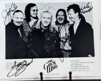 Black-and-white promotional photograph of The Guess Who, measuring 8” H x 10” W. Features five signatures on the front. Includes names of former members and band details at the bottom of the image. Housed in a protective sleeve.