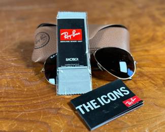 Ray-Ban Aviator sunglasses featuring gold-tone frames and dark lenses with visible Ray-Ban branding on the lens. Includes a brown leather-like protective case with the Ray-Ban logo, cleaning cloth, and ‘The Icons’ booklet. All items are in excellent condition and appear unused.