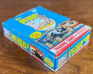 A box of Topps Desert Storm trading cards and stickers from the early 1990s. The box contains 36 sealed packs, each featuring 8 trading cards and 1 sticker, as indicated on the packaging. The box shows visible wear but remains structurally sound, and the packs inside are factory-sealed. Collectible item reflecting historical themes showcased in trading cards.