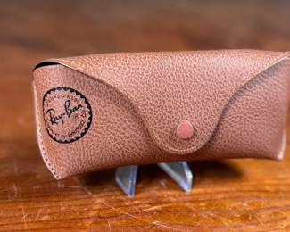 Ray-Ban Aviator sunglasses featuring gold-tone frames and dark lenses with visible Ray-Ban branding on the lens. Includes a brown leather-like protective case with the Ray-Ban logo, cleaning cloth, and ‘The Icons’ booklet. All items are in excellent condition and appear unused.