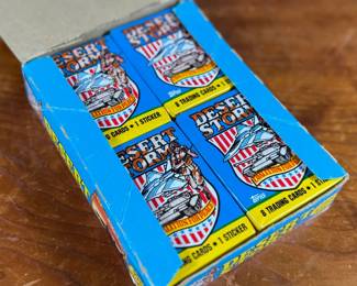 A box of Topps Desert Storm trading cards and stickers from the early 1990s. The box contains 36 sealed packs, each featuring 8 trading cards and 1 sticker, as indicated on the packaging. The box shows visible wear but remains structurally sound, and the packs inside are factory-sealed. Collectible item reflecting historical themes showcased in trading cards.
