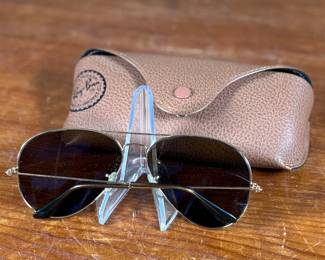 Ray-Ban Aviator sunglasses featuring gold-tone frames and dark lenses with visible Ray-Ban branding on the lens. Includes a brown leather-like protective case with the Ray-Ban logo, cleaning cloth, and ‘The Icons’ booklet. All items are in excellent condition and appear unused.