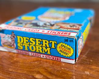 A box of Topps Desert Storm trading cards and stickers from the early 1990s. The box contains 36 sealed packs, each featuring 8 trading cards and 1 sticker, as indicated on the packaging. The box shows visible wear but remains structurally sound, and the packs inside are factory-sealed. Collectible item reflecting historical themes showcased in trading cards.