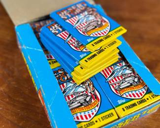 A box of Topps Desert Storm trading cards and stickers from the early 1990s. The box contains 36 sealed packs, each featuring 8 trading cards and 1 sticker, as indicated on the packaging. The box shows visible wear but remains structurally sound, and the packs inside are factory-sealed. Collectible item reflecting historical themes showcased in trading cards.