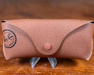 Ray-Ban Aviator sunglasses featuring gold-tone frames and dark lenses with visible Ray-Ban branding on the lens. Includes a brown leather-like protective case with the Ray-Ban logo, cleaning cloth, and ‘The Icons’ booklet. All items are in excellent condition and appear unused.