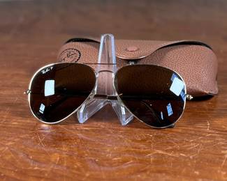 Ray-Ban Aviator sunglasses featuring gold-tone frames and dark lenses with visible Ray-Ban branding on the lens. Includes a brown leather-like protective case with the Ray-Ban logo, cleaning cloth, and ‘The Icons’ booklet. All items are in excellent condition and appear unused.