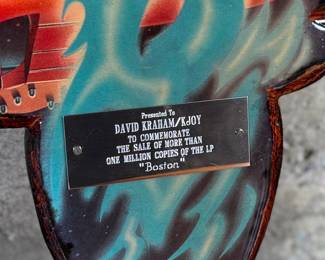 Boston commemorative plaque celebrating the sale of more than one million copies of the LP “Boston.” The design replicates the iconic album cover, featuring a UFO motif with vibrant colors and a mounted metal dedication plate. Includes documentation verifying authenticity. Constructed from wood and metal materials, with visible mounting hardware on the reverse side. Accompanied with a certificate of authenticity.