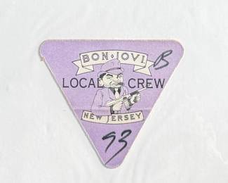 A triangular Bon Jovi Local Crew pass from 1993, featuring the text “Bon Jovi Local Crew” alongside “New Jersey” and an illustration of a suited cartoon figure holding weapons. Marked with the number “93” and a handwritten “B.” Made of adhesive-backed material.