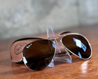Ray-Ban Aviator sunglasses featuring gold-tone frames and dark lenses with visible Ray-Ban branding on the lens. Includes a brown leather-like protective case with the Ray-Ban logo, cleaning cloth, and ‘The Icons’ booklet. All items are in excellent condition and appear unused.