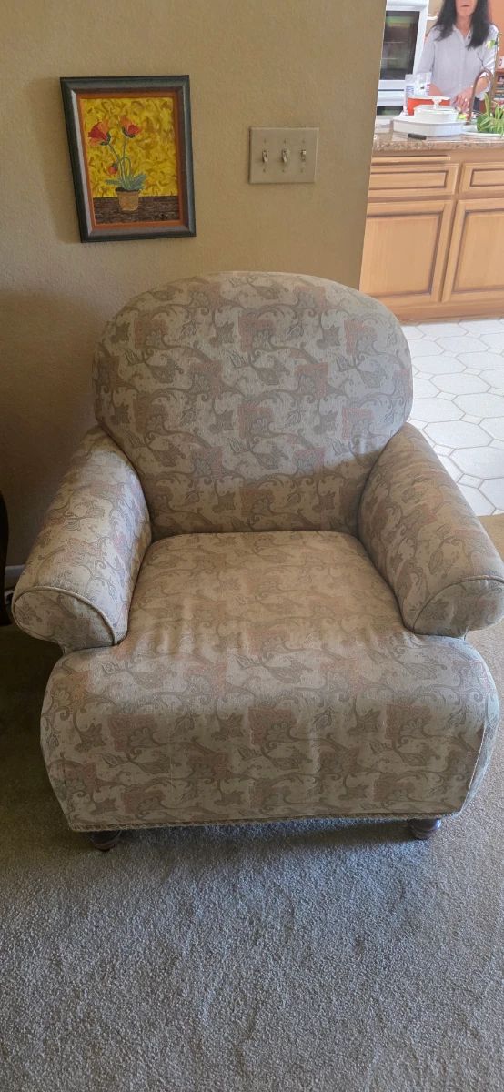 Upholstered club chair with a soft, rounded back and rolled arms in a traditional style fabric featuring a subtle paisley/leaf pattern in muted beige and rose tones. The seat is wide and deep with a low profile, and the chair sits on short turned wooden feet, giving it a comfortable, classic living‑room look.