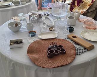 White porcelain bowls, plates, cups, and saucers alongside glassware, a clear punch bowl, wooden serving tools, rattan napkin rings, and decorative items.