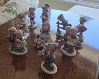 9 Hummel figurines depicting children in traditional German folk costumes engaged in everyday activities like playing music, carrying baskets, and farm work