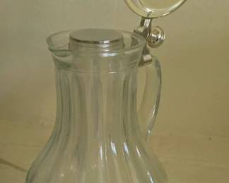 Vintage ice water pitcher featuring ribbed clear glass body designed to hold ice in an inner compartment, topped by a hinged silver-plated lid and handle with a thumb flip for pouring.
