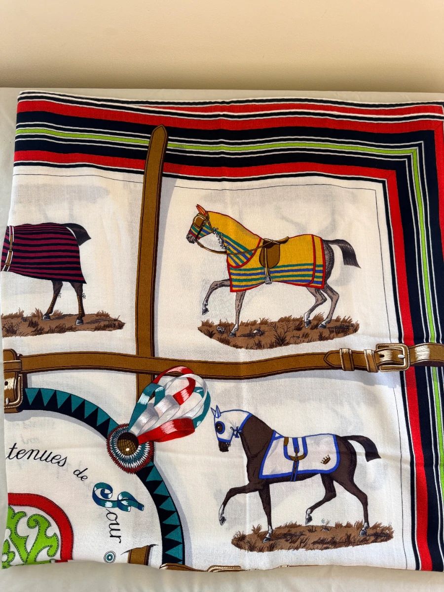 Hermès cashmere large scarf titled "Couvertures et Tenues de Jour" (translated as "Blankets and Day Wear"). Hermès Hermès +1 Designed by artist Jacques Eudel and first issued in 1974, this design is a staple of the Hermès equestrian heritage. The motif features ten horses, each fitted with a different style of blanket or "outfit" required for various equestrian activities, elegantly framed by leather bridles