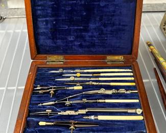 An antique drafting instrument set, featuring various compasses, ruling pens, and dividers for technical drawing, housed within a wooden case lined with blue velvet