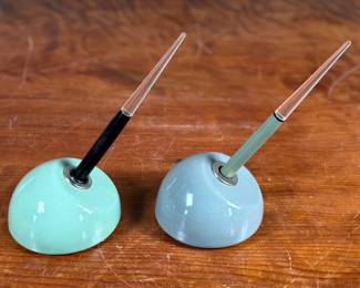 Esterbrook dip pens featuring metal nibs marked ‘Esterbrook 9556’ with black and green handles. Each pen is paired with a ceramic holder in complementing colors: green and blue-gray. Both pens are marked ‘Esterbrook Made in U.S.A.’ on the handles, denoting their authenticity. Well-preserved and functional writing tools, ideal for collectors or calligraphy enthusiasts.