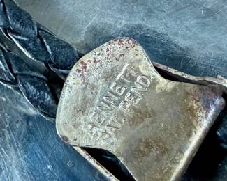 Silver bolo tie featuring a decorative oval slide adorned with turquoise and coral cabochons set in an intricate feather motif. The tie features a black braided leather cord with silver-tone tips and is marked ‘Bennett Pat Pend’ on the back. Shows slight age-related wear on metal surfaces consistent with use.