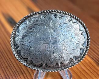 Sterling silver belt buckle crafted by Kustom Kraft featuring an intricate engraved pattern with foliate and scroll motifs. The oval-shaped buckle is bordered with a twisted rope-style edge. The back is stamped with ‘STERLING’ and the Kustom Kraft hallmark. Measures, 3” H x 4” W with a total approx. weight of 87.00 grams.